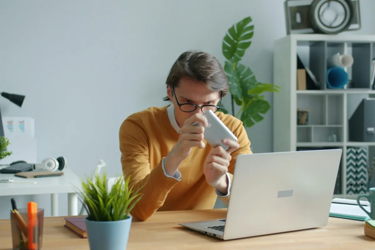 A man at a desk peering at his phone instead of his open laptop