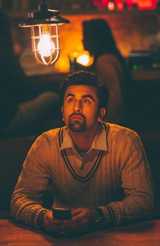 Ranbir Kapoor as Ved in Tamasha, sitting alone at a café table beneath a hanging lamp, looking upward