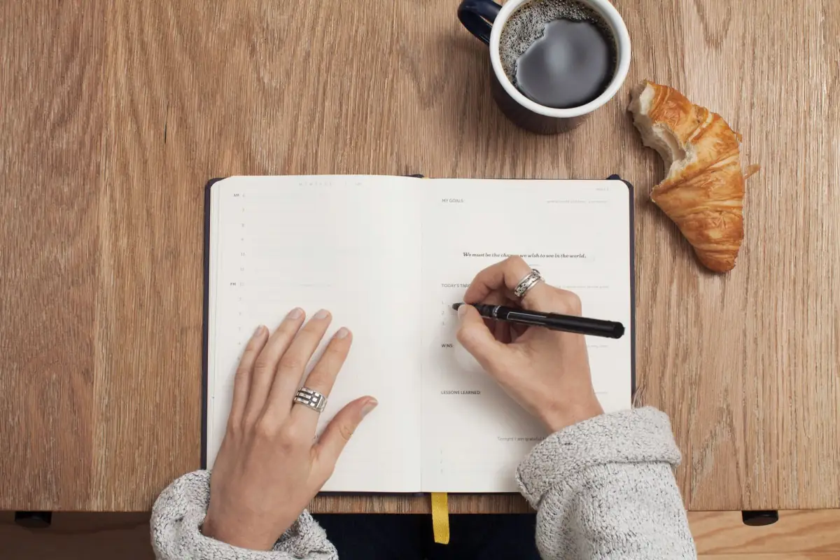 Someone writing in a planner at a wooden table, with coffee and a croissant