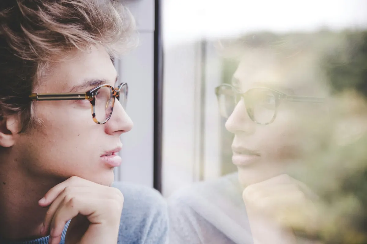 A young man with glasses gazing out of a window, his reflection in the glass