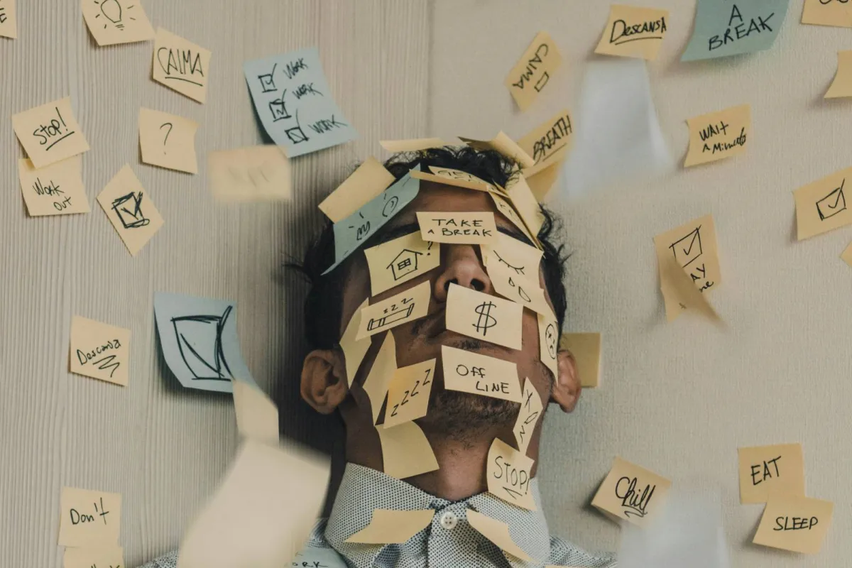 A man leaning back against a wall, his face and shirt covered in sticky notes reading “take a break”, “stop”, “chill” and “sleep”