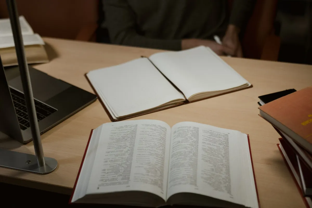 A study desk with an open book, a blank notebook, a laptop and a stack of books