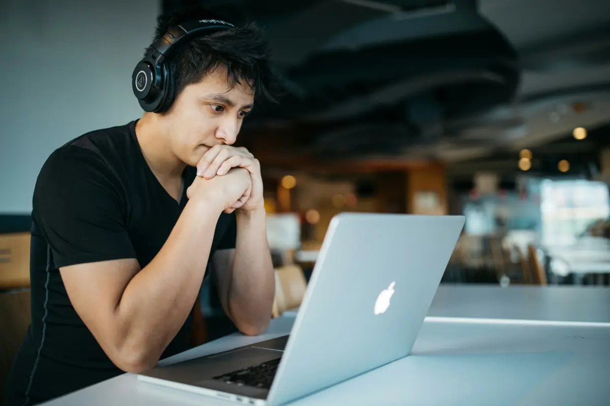 A young man with headphones, hands clasped, looking thoughtfully at his laptop