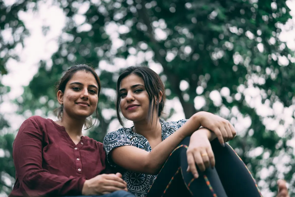 Two young Indian women sitting together outdoors under trees, smiling